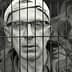 A man wearing glasses and a cap, looking through metal bars with a serious expression. - Olive Oil Times