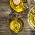 Glass bowl and bottle containing olive oil with herbs on a wooden surface. - Olive Oil Times