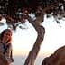 Woman with curly hair sitting on a branch of an olive tree during sunset. - Olive Oil Times