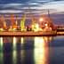 Cargo ships docked at a port with cranes illuminated by lights during twilight hours. - Olive Oil Times