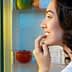 A woman with dark hair smiling while looking into an open refrigerator filled with food items. - Olive Oil Times