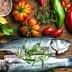 Two whole fish placed on a wooden cutting board surrounded by various vegetables and herbs. - Olive Oil Times