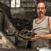 Man in a tank top holding a bowl of charcoal in a dimly lit room with charcoal piles. - Olive Oil Times