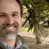 Man with a beard smiling in an olive grove with olive branches in the background. - Olive Oil Times
