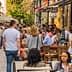 Crowd of people dining at outdoor tables along a city street with pedestrians walking by. - Olive Oil Times