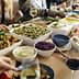 A dining table filled with various dishes, including salads, meats, and side dishes, with people dining around it. - Olive Oil Times
