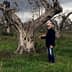 A man in a black jacket standing next to an olive tree in a field. - Olive Oil Times