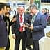 Four individuals engaged in conversation while examining olive oil products at an event. - Olive Oil Times