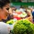 A woman looking at fresh vegetables in a market, with various greens and produce visible in the background. - Olive Oil Times