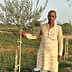 Man in traditional attire standing beside a young olive tree in a field. - Olive Oil Times