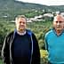 Two men standing side by side in an olive grove with a hillside village in the background. - Olive Oil Times