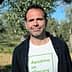 Man standing in an olive grove wearing a white t-shirt with green text. - Olive Oil Times