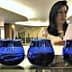 Four blue glass containers arranged on a table with a woman reviewing documents in the background. - Olive Oil Times