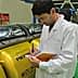 Man in a lab coat taking notes while observing yellow machinery in a facility. - Olive Oil Times