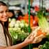 A woman smiling while holding a piece of ginger in a market filled with fresh produce. - Olive Oil Times