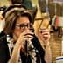 Woman holding a small cup while tasting olive oil at a dining table with flags in the background. - Olive Oil Times