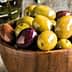 A wooden bowl filled with various types of olives including green and black varieties. - Olive Oil Times