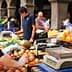 A vendor weighing apples at a market while another vendor assists a customer in the background. - Olive Oil Times