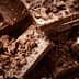 Pieces of dark chocolate stacked with chocolate shavings scattered around on a wooden surface. - Olive Oil Times