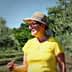 Woman wearing a yellow shirt and hat standing among olive trees in an olive grove. - Olive Oil Times