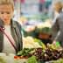 Woman examining fresh vegetables at a market stall surrounded by other shoppers. - Olive Oil Times