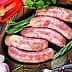 Five raw sausages arranged on a black platter with herbs and garlic nearby. - Olive Oil Times