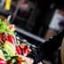 Woman with a scarf selecting red and green peppers at a market stand filled with vegetables. - Olive Oil Times