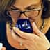Woman with glasses smelling olive oil from a blue tasting cup while holding a small container. - Olive Oil Times