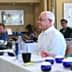 Man with white hair seated at a conference table with participants in the background during a meeting. - Olive Oil Times