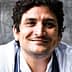 Smiling chef with curly hair wearing a white chef jacket and a gray apron. - Olive Oil Times