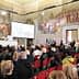A large audience seated in a historical hall with elaborate murals on the walls. - Olive Oil Times