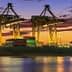 Cargo ship docked at a port with cranes and containers during sunset with colorful sky. - Olive Oil Times