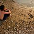 A child sitting on dry, cracked earth near a small body of water. - Olive Oil Times