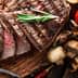 Sliced grilled steak on a wooden cutting board with tomatoes, mushrooms, and sauce. - Olive Oil Times