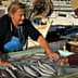A woman in an apron selling fish at a market while a man points at the fish on display. - Olive Oil Times