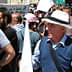 An older man wearing a white hat and holding a small flag at a crowded public event. - Olive Oil Times