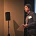 A man presenting at a conference with a microphone and projector screen in the background. - Olive Oil Times