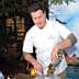 Chef in a white uniform mixing ingredients in a bowl at an outdoor event with a backdrop. - Olive Oil Times