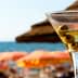 A martini glass filled with a clear liquid and three green olives, with a beach background. - Olive Oil Times