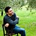 Man with dark hair sitting on a wooden chair, smiling in an olive grove. - Olive Oil Times