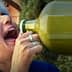 Woman with gray hair drinking from a large green glass bottle outdoors. - Olive Oil Times