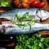 Two raw fish placed on a wooden cutting board surrounded by fresh vegetables and herbs. - Olive Oil Times