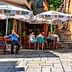 Outdoor café scene featuring patrons seated under large umbrellas in a narrow street. - Olive Oil Times