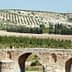 A stone bridge with three arches spanning over a river, surrounded by olive trees on a hillside. - Olive Oil Times