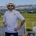 Chef wearing a white coat, hat, and sunglasses standing outdoors with a scenic background. - Olive Oil Times