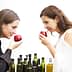 Two women holding red tasting cups while smelling olive oil from bottles in front of them. - Olive Oil Times