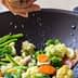 Person sprinkling salt over a mix of vegetables in a black wok with a wooden spoon. - Olive Oil Times