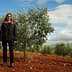 Individual standing in an olive grove with green trees and red soil in the background. - Olive Oil Times