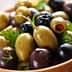 A wooden bowl filled with a variety of green and black olives, garnished with herbs. - Olive Oil Times