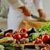 A variety of fresh vegetables, herbs, and cooking ingredients arranged on a kitchen counter with a chef preparing food in the background. - Olive Oil Times
